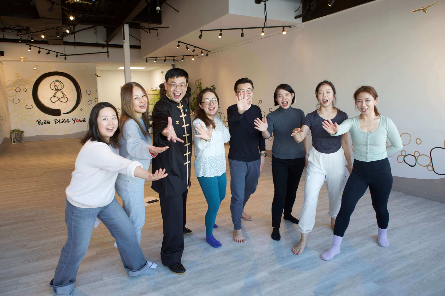 Group of eight adults smiling and reaching toward the camera in a bright yoga studio, with the “Pure Bliss Yoga” logo on the back wall.